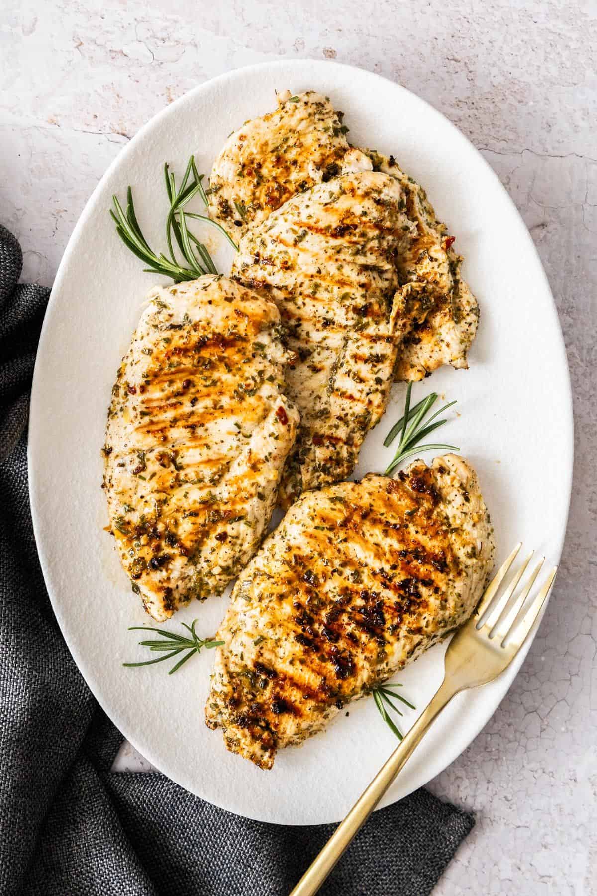 White oval platter of Marinated Chicken Breast on a grey cloth, plate garnished with some rosemary, with a fork on the side.