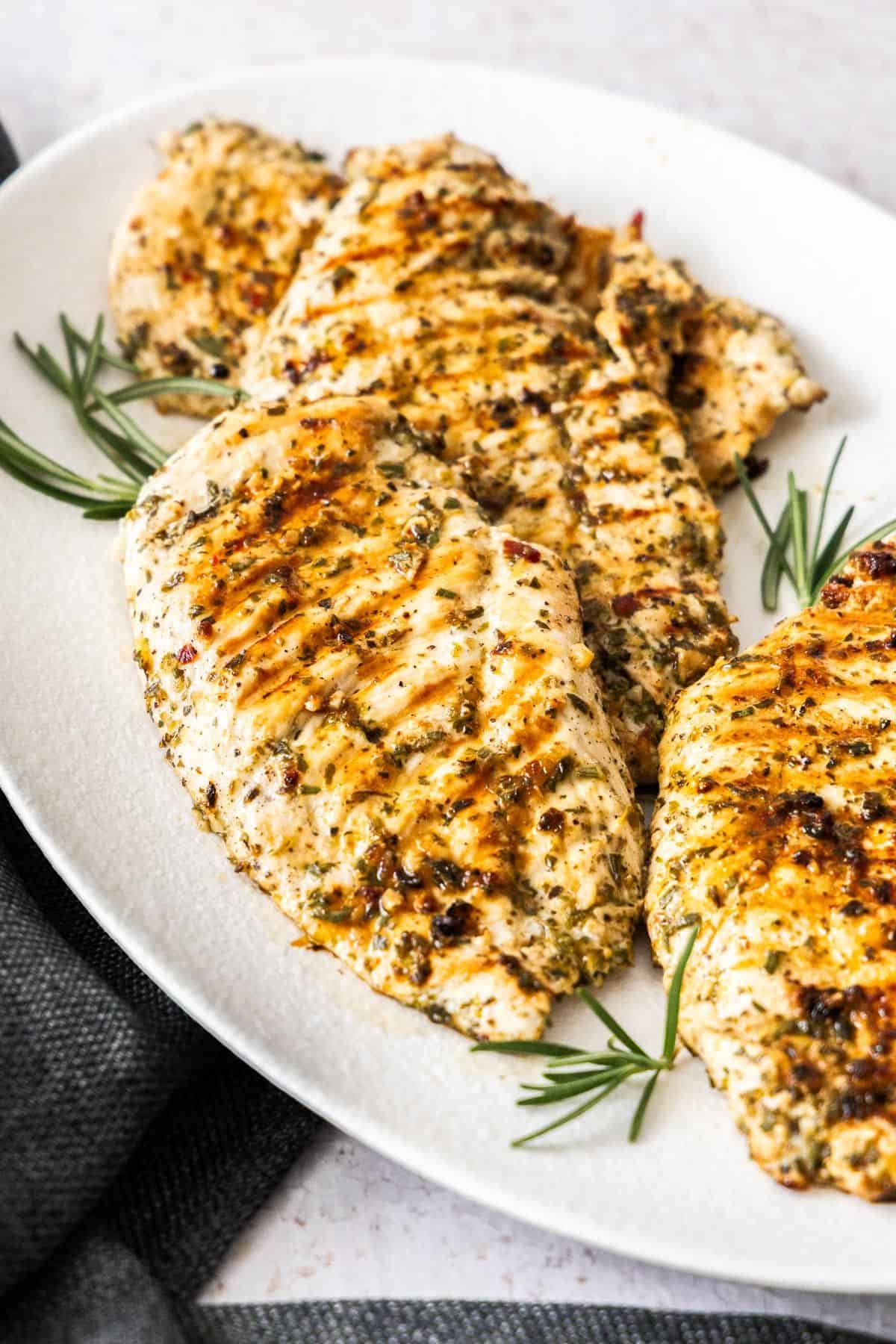 White oval platter of Marinated Chicken Breast on a grey cloth, plate garnished with some rosemary.