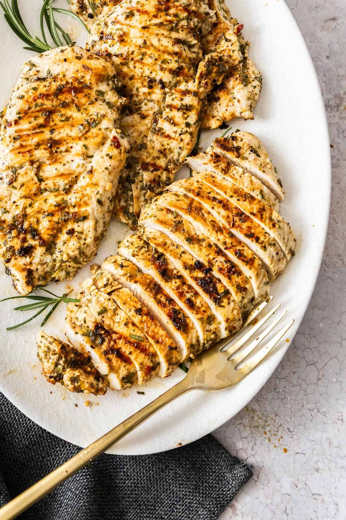 White oval platter of Marinated Chicken Breast on a grey cloth, plate garnished with some rosemary, with a fork on the side.