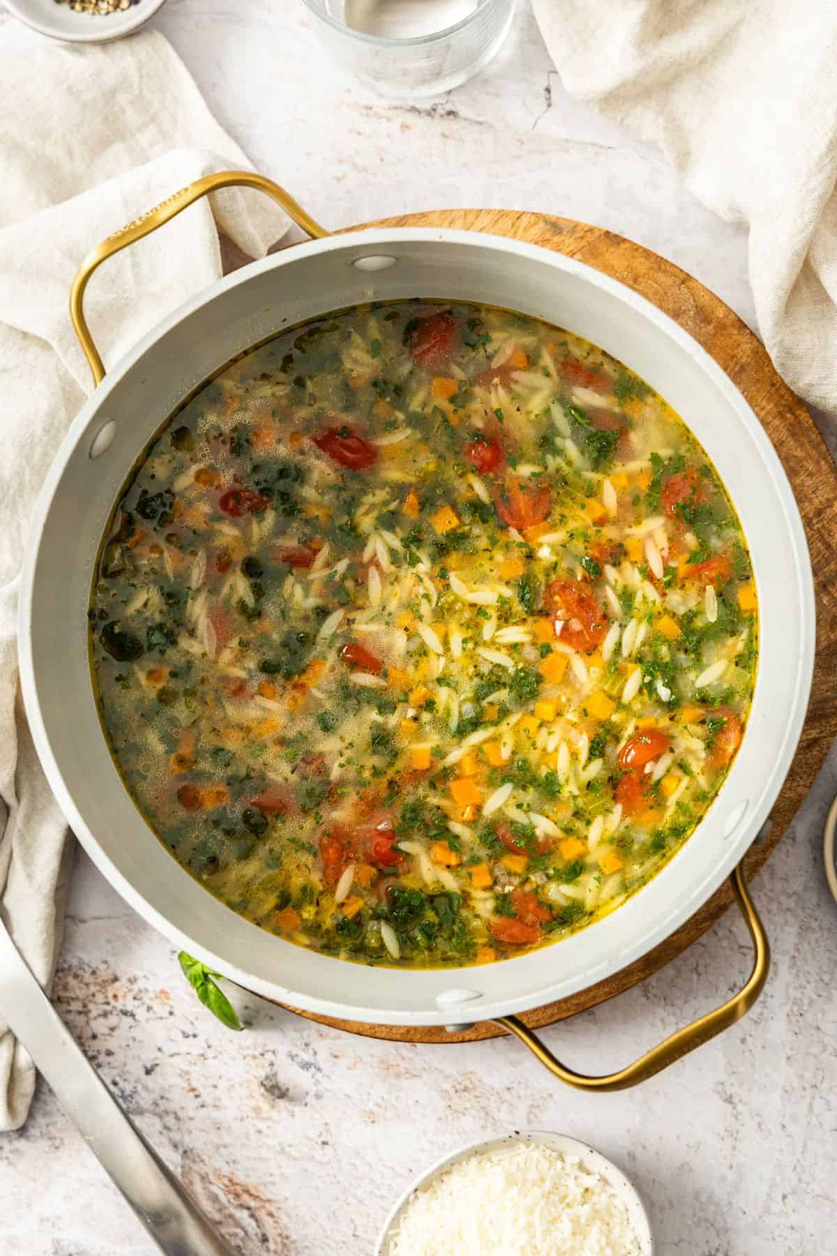 Large saucepan of Mediterranean Orzo Soup, sitting on a round wooden board.