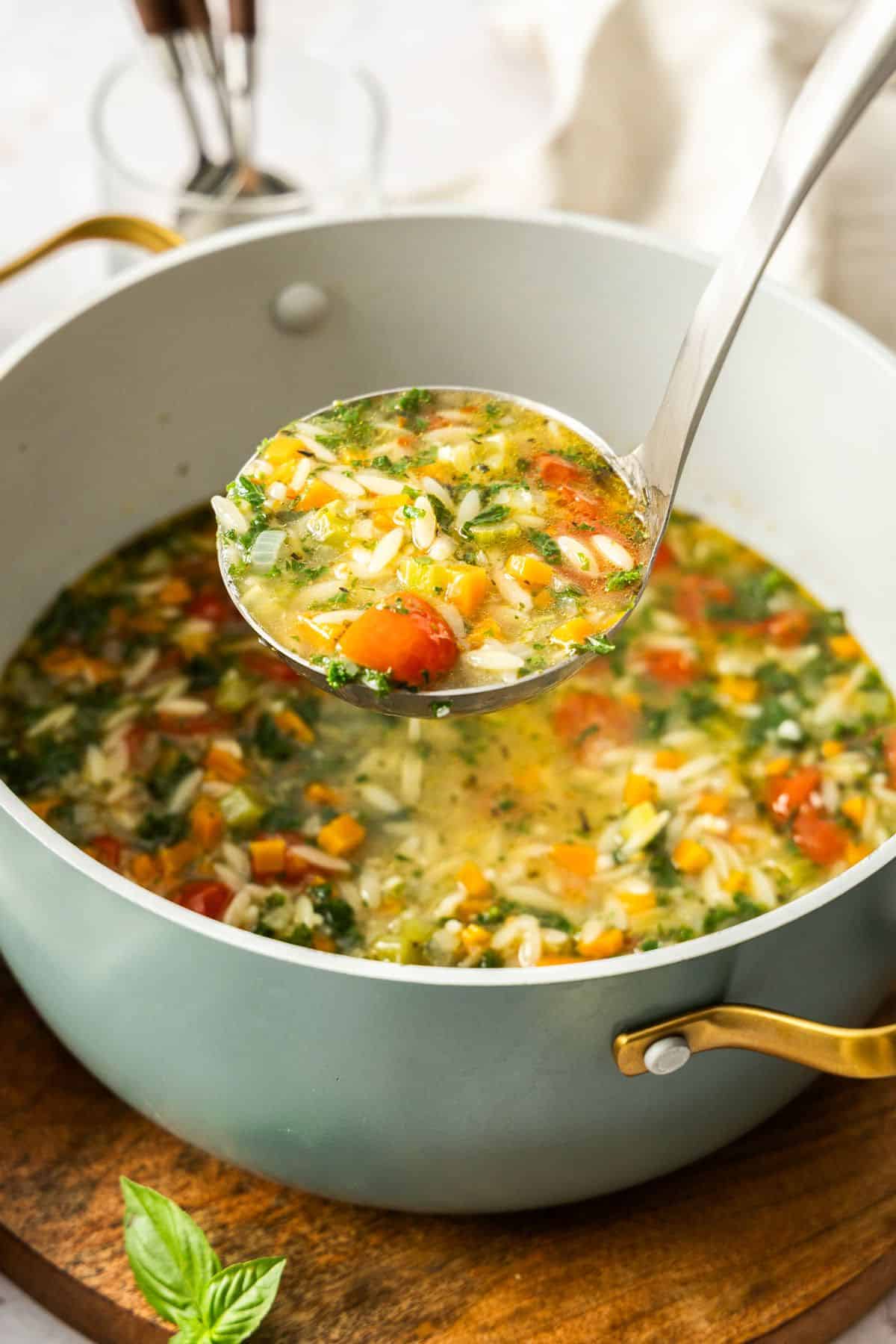Large saucepan of Mediterranean Orzo Soup, sitting on a round wooden board, with a ladle lifting up some soup.