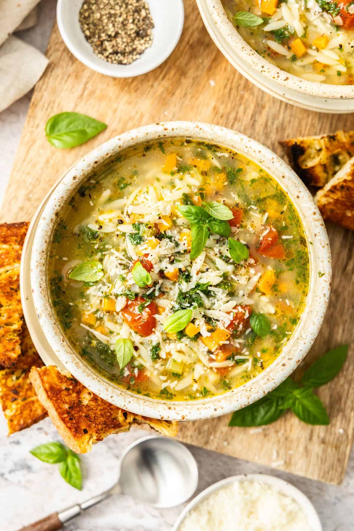 Two round bowls of Mediterranean Orzo Soup, garnished with Parmesan and Basil, with some toasted bread on the edge.