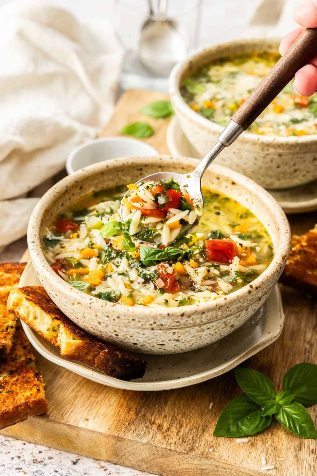 Two round bowls of Mediterranean Orzo Soup, one with a spoon lifting up some soup, with some toasted bread on the edge.