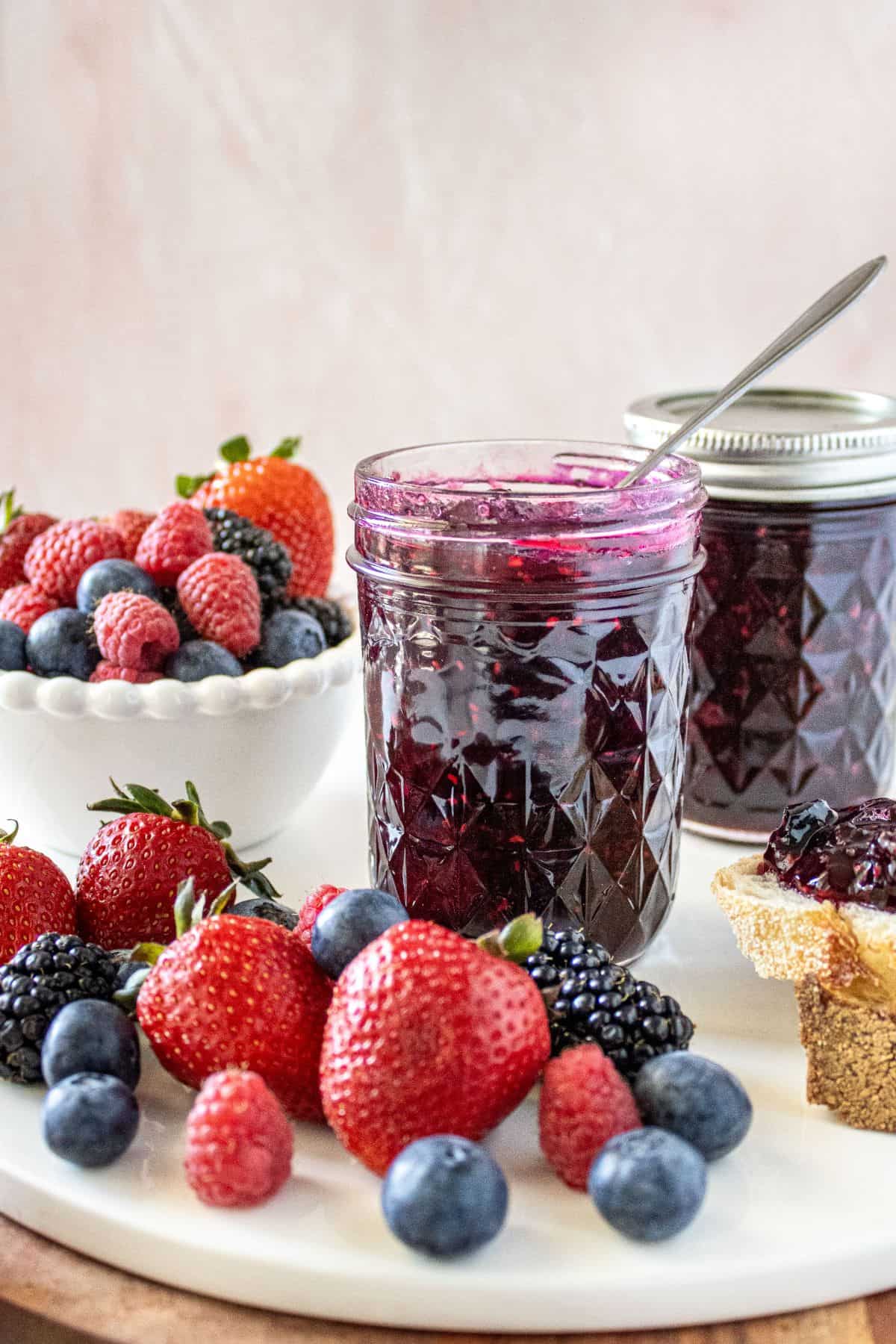 Jar of Mixed Berry Jam with a spoon in it, sitting on a marble plate with some fresh berries around the edge.