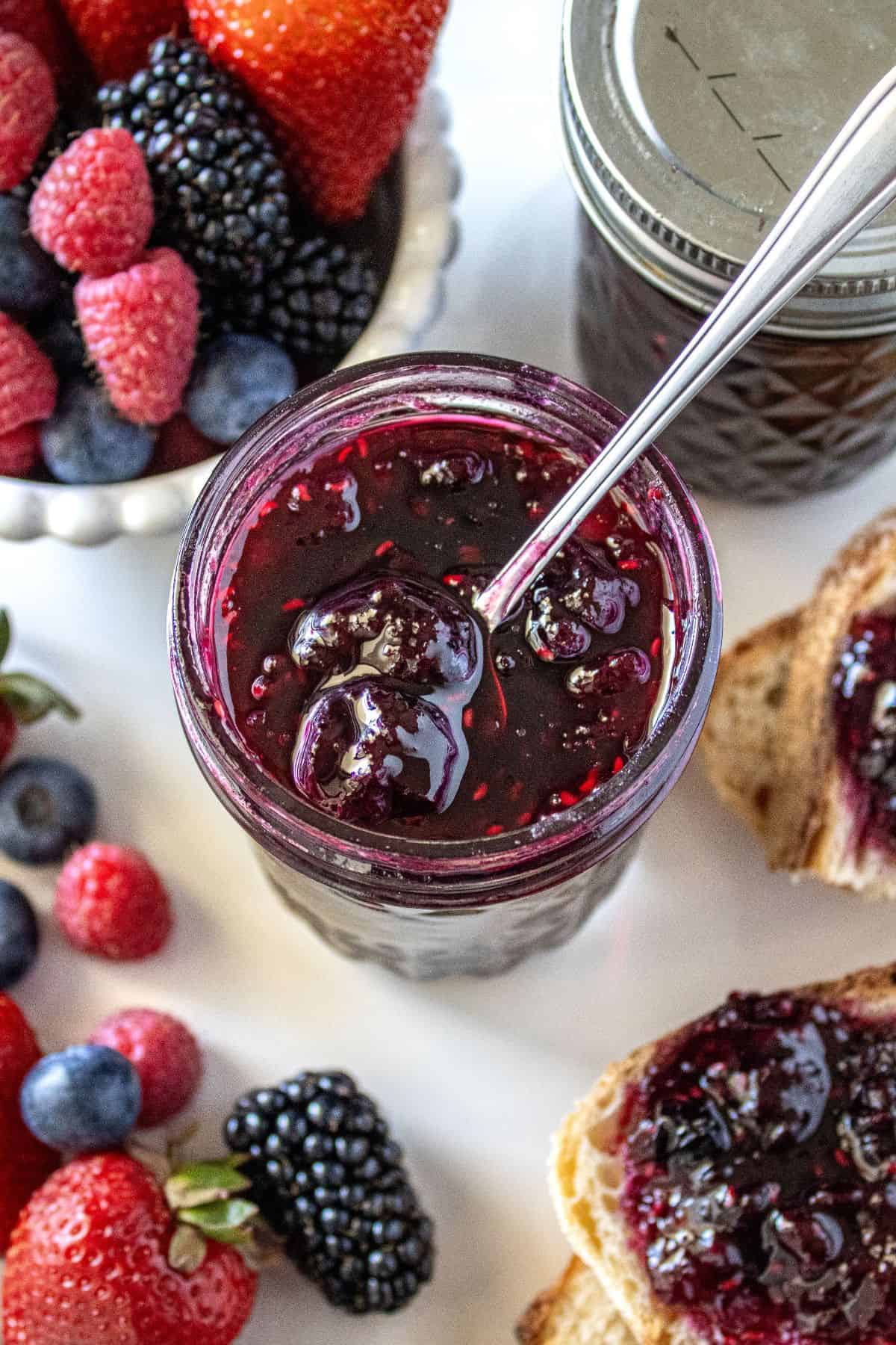 Jar of Mixed Berry Jam with a spoon in it, sitting on a marble plate with some fresh berries around the edge.