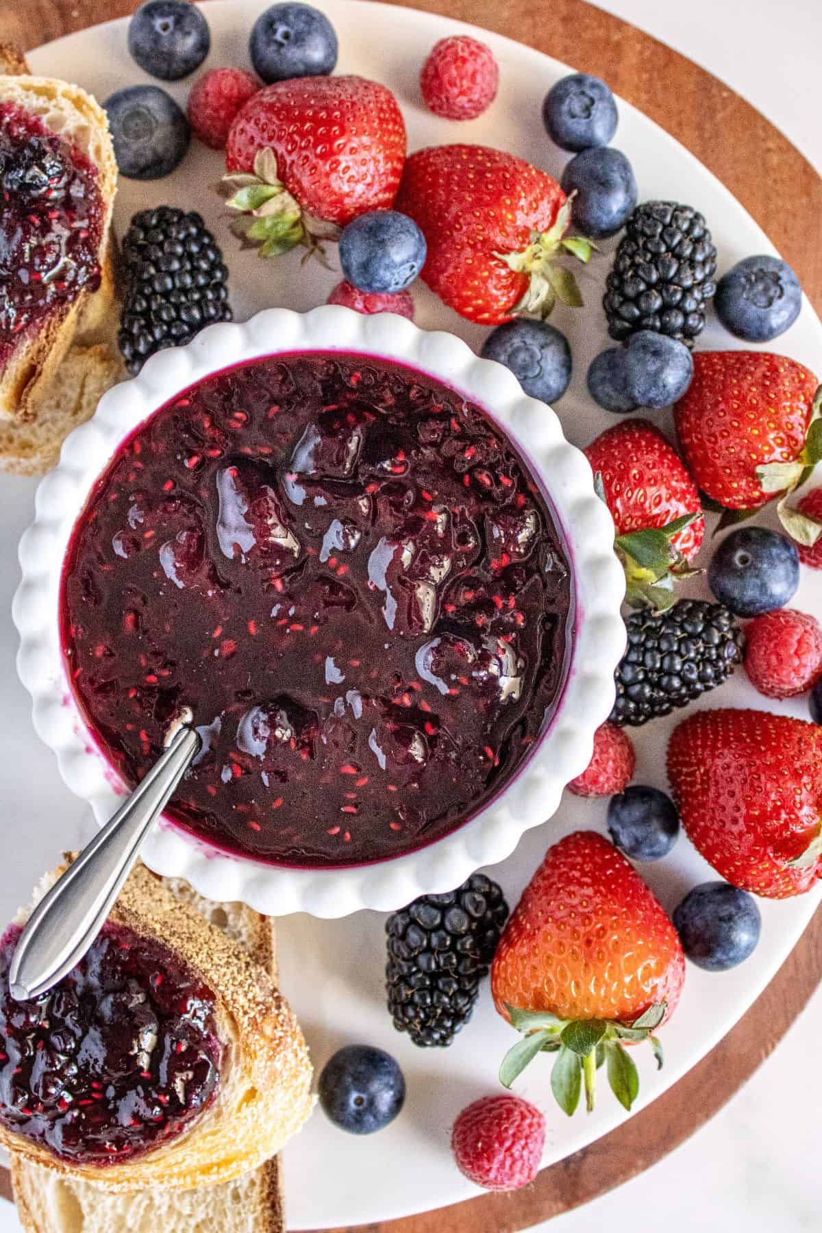 Small white dish of Mixed Berry Jam on a marble plate with some fresh berries and bread around the edge.