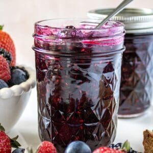 Jar of Mixed Berry Jam with a spoon in it, sitting on a marble plate with some fresh berries around the edge.