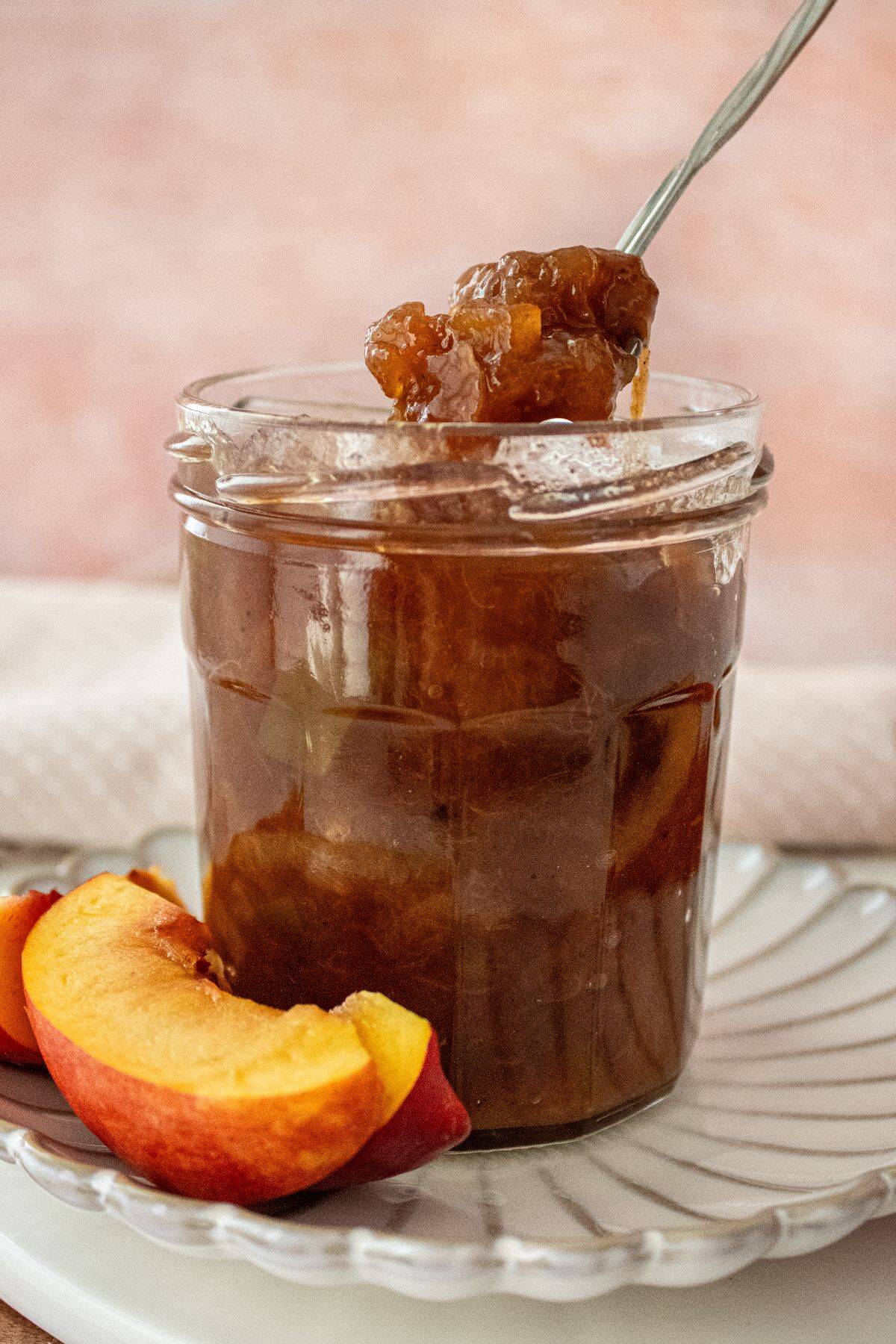 Open glass jar of Nectarine Chutney, sitting on a small white plate, with a spoon scooping up some chutney.