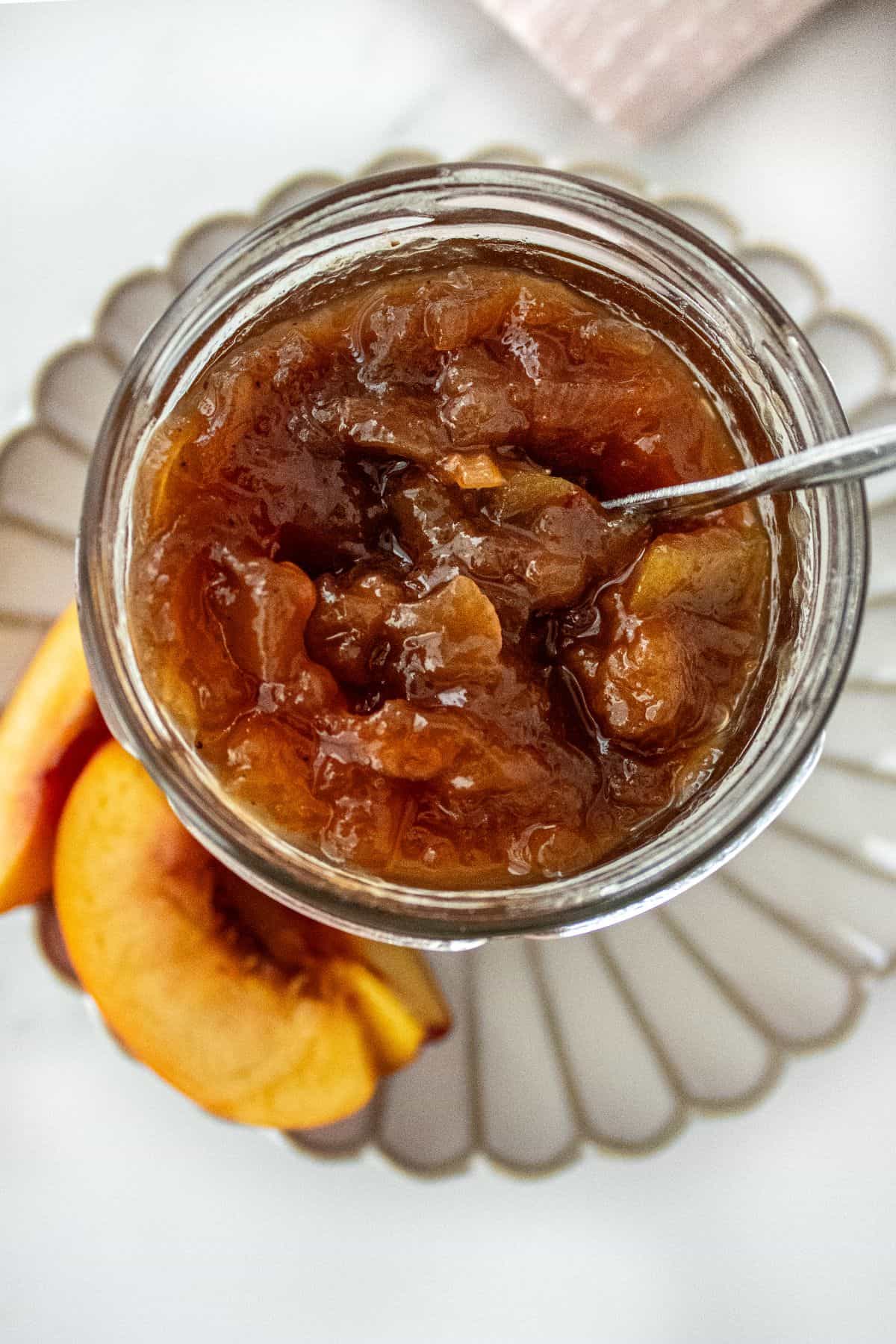 Open glass jar of Nectarine Chutney, sitting on a small white plate, with a spoon sitting in the jar.
