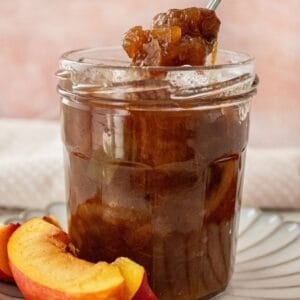 Open glass jar of Nectarine Chutney, sitting on a small white plate, with a spoon scooping up some chutney.