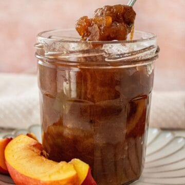 Open glass jar of Nectarine Chutney, sitting on a small white plate, with a spoon scooping up some chutney.