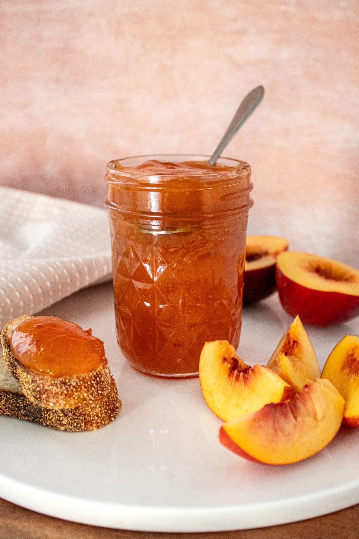 Open jar of Nectarine Jam on a white plate, with some pieces of nectarine and bread around the edge.