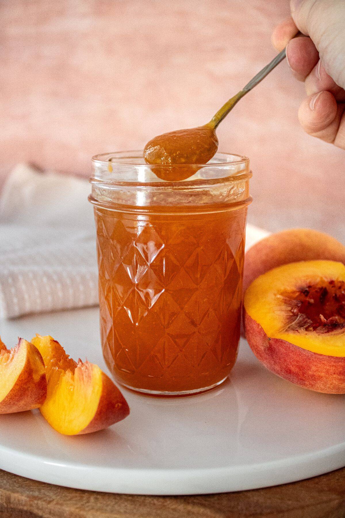 Open glass jar of Peach Jam sitting on a white plate, with a spoon scooping up some jam.