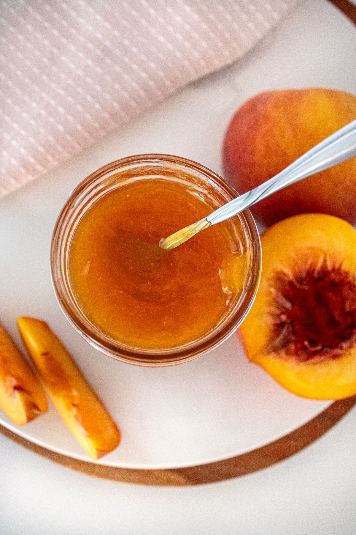 Open glass jar of Peach Jam sitting on a white plate, with a spoon sitting in the jar, and some peaches around the edge.