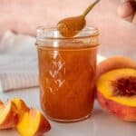 Open glass jar of Peach Jam sitting on a white plate, with a spoon scooping up some jam.