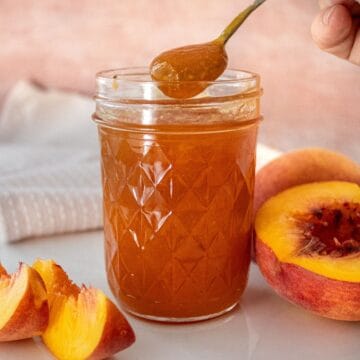 Open glass jar of Peach Jam sitting on a white plate, with a spoon scooping up some jam.