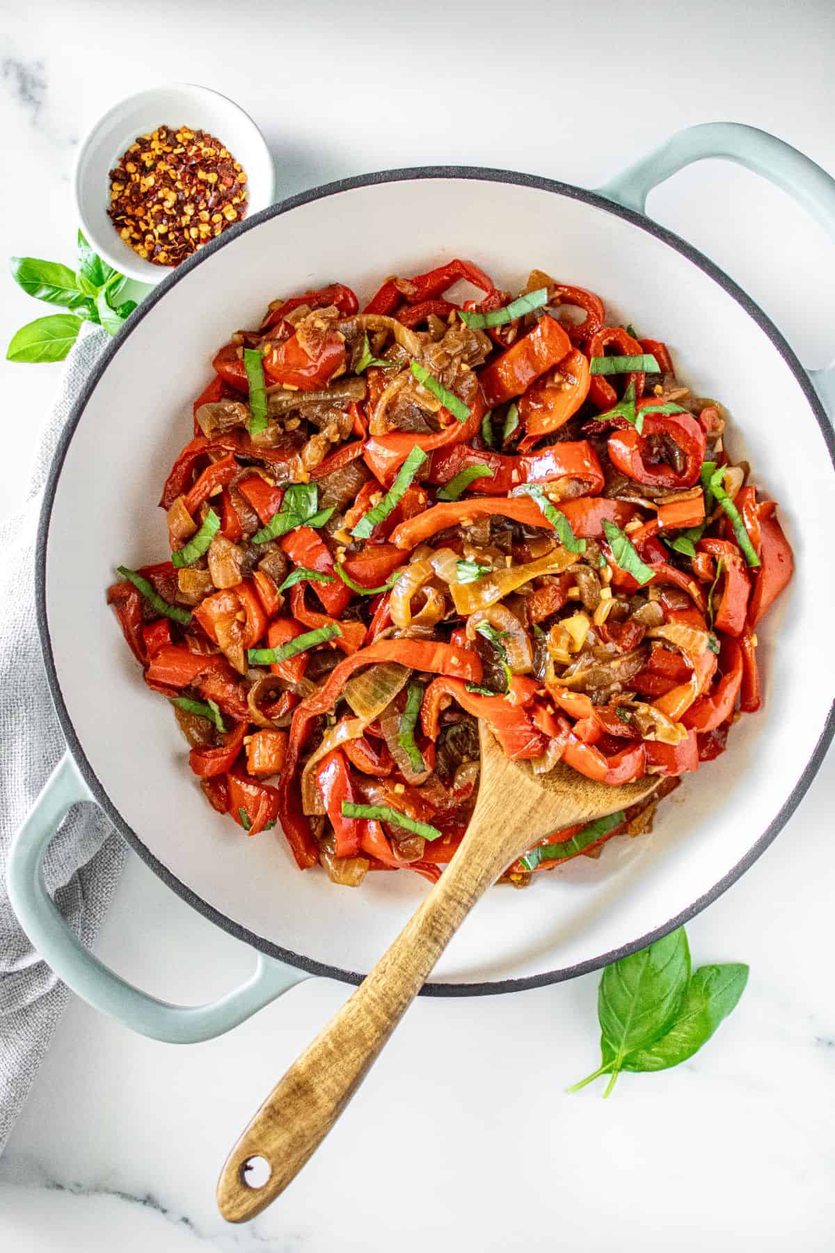 Sautéed Peppers and Onions in a large ceramic dish with a wooden spoon, and some basil leaves and chilli around the edge.