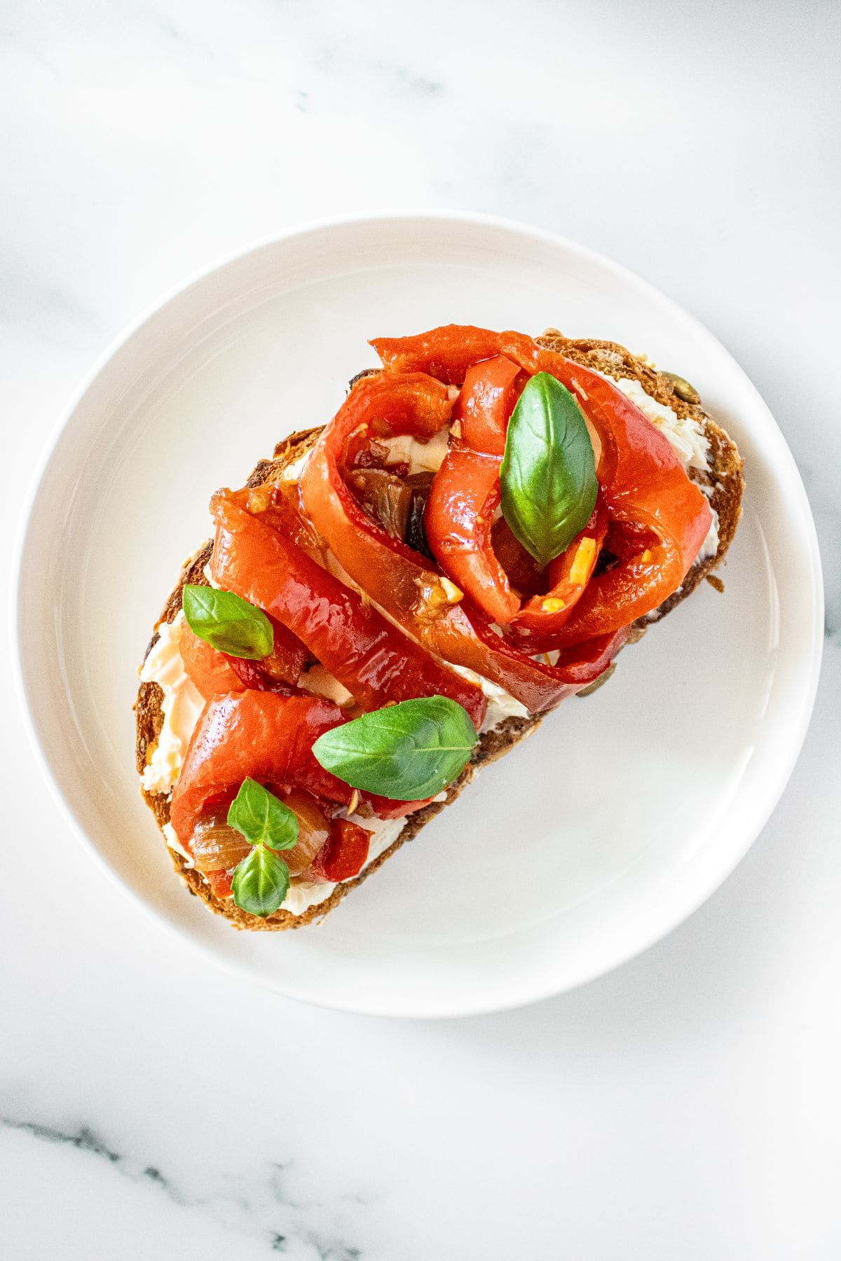 A small white plate with a slice of bread, topped with cream cheese and some Sautéed Peppers and Onions and basil leaves.