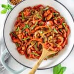 Sautéed Peppers and Onions in a large ceramic dish with a wooden spoon, and some basil leaves and chilli around the edge.