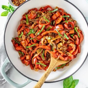 Sautéed Peppers and Onions in a large ceramic dish with a wooden spoon, and some basil leaves and chilli around the edge.
