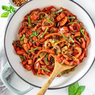 Sautéed Peppers and Onions in a large ceramic dish with a wooden spoon, and some basil leaves and chilli around the edge.
