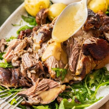 Platter with Slow Roasted Pork Shoulder being drizzled with gravy, with some rocket leaves and potatoes around the edge.