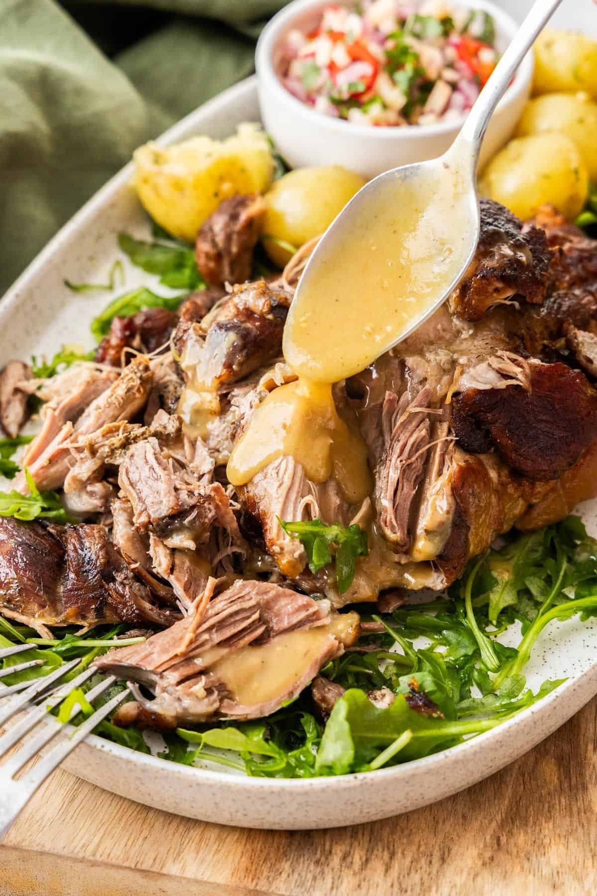 Platter with Slow Roasted Pork Shoulder being drizzled with gravy, with some rocket leaves and potatoes around the edge.