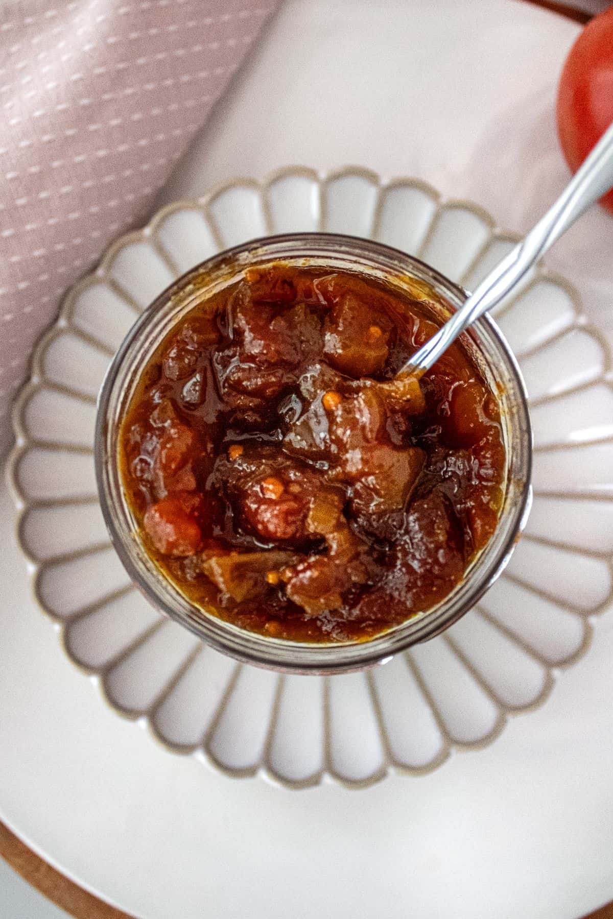 Jar of Tomato Chutney, on a round white plate, with a spoon in the jar.