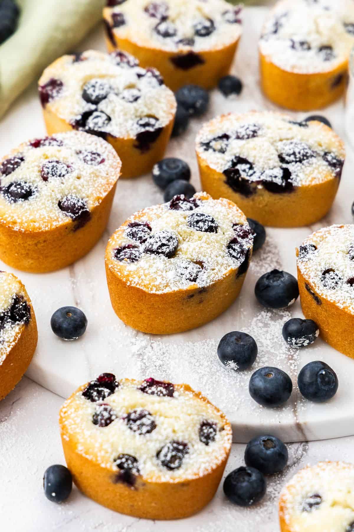 Gluten-Free Blueberry Friands on a round marble plate, dusted with icing/powdered sugar, with some blueberries scattered.