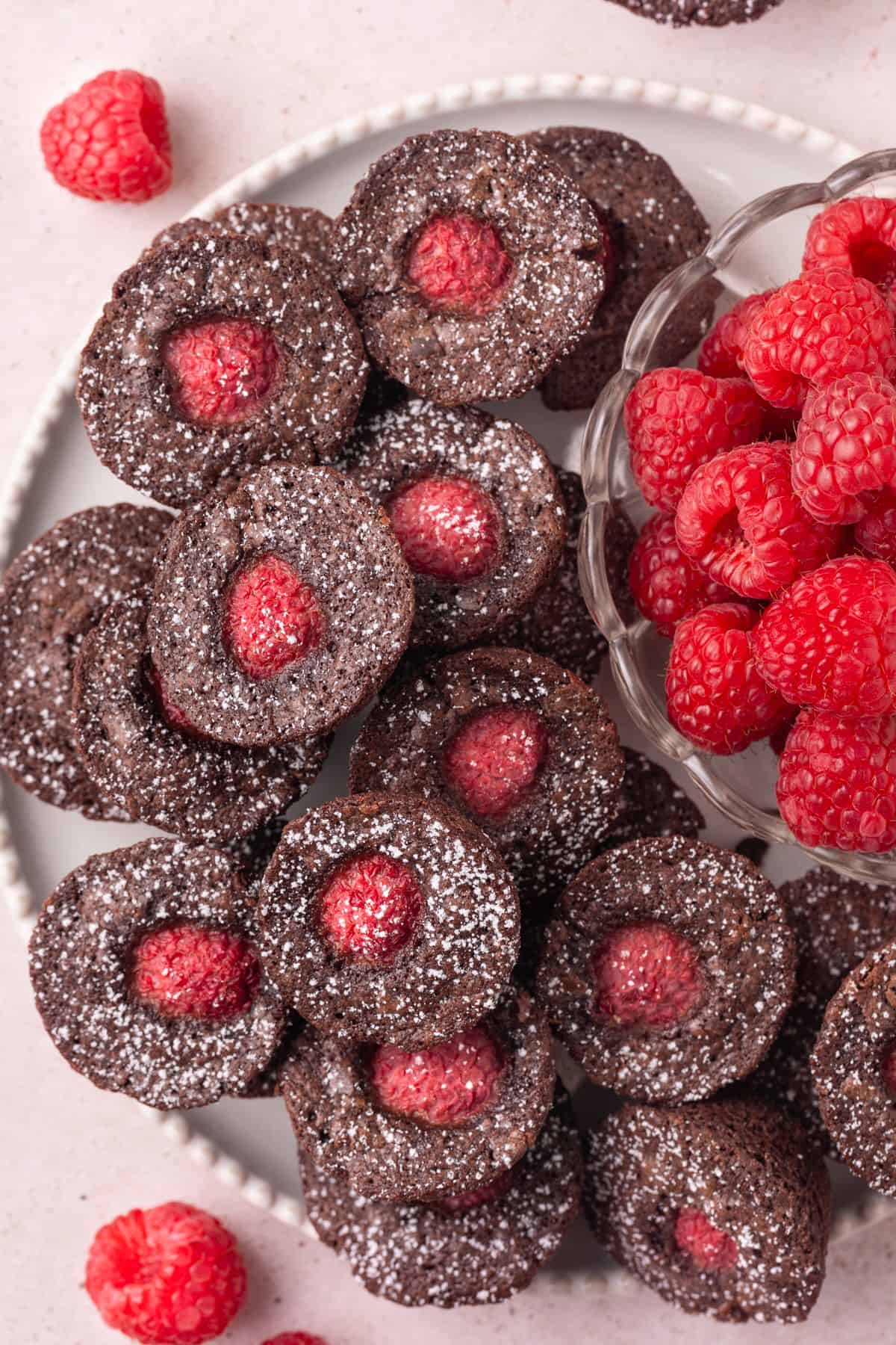 Round white plate with Chocolate Raspberry Bites dusted with icing/powdered sugar, and a small bowl of fresh raspberries.