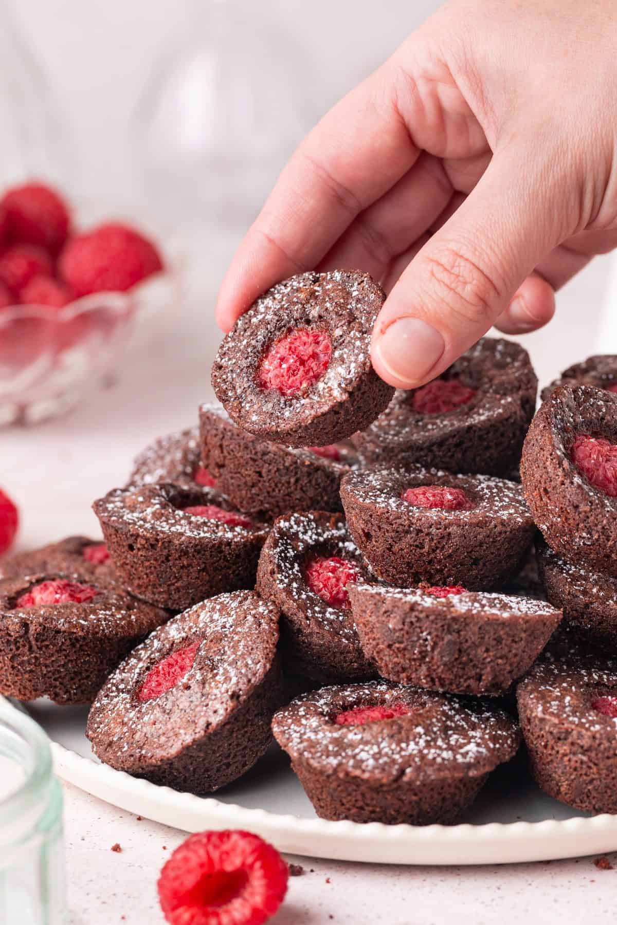 Round white plate with Chocolate Raspberry Bites dusted with icing/powdered sugar, with a hand picking up one of the bites.