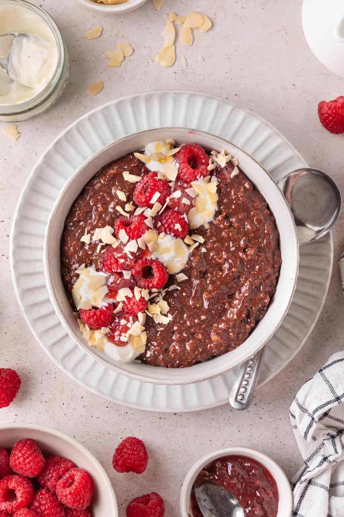 Round bowl of Chocolate Raspberry Porridge topped with yoghurt, flaked almonds and raspberries, with a spoon on the edge.