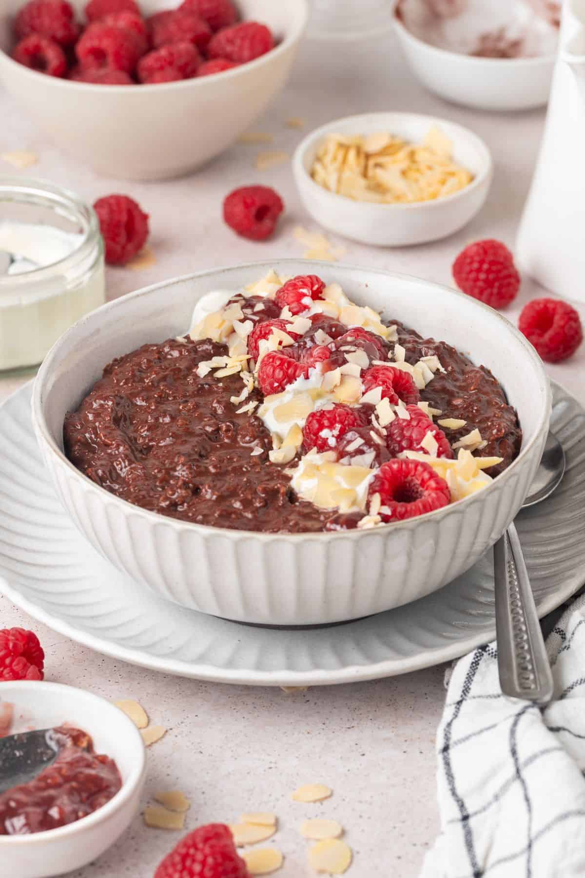Round bowl of Chocolate Raspberry Porridge topped with yoghurt, flaked almonds and raspberries, with a spoon on the edge.