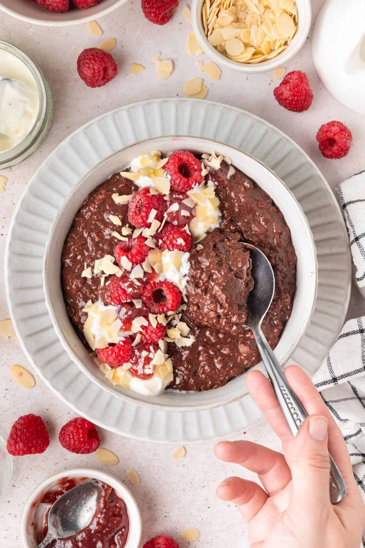 Spoon dipping in to a round bowl of Chocolate Raspberry Porridge topped with yoghurt, flaked almonds and raspberries.
