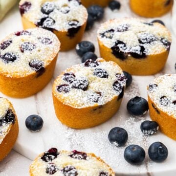 Gluten-Free Blueberry Friands on a round marble plate, dusted with icing/powdered sugar, with some blueberries scattered.
