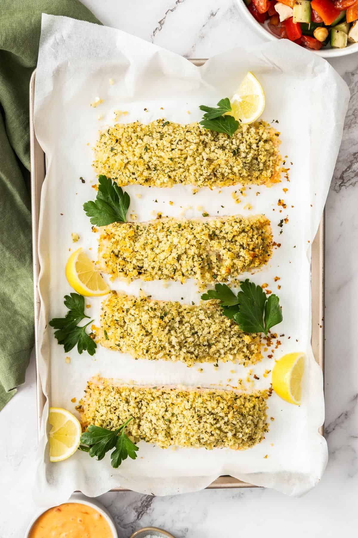 Baking tray with cooked Herb Panko Salmon, garnished with some parsley leaves and lemon wedges.