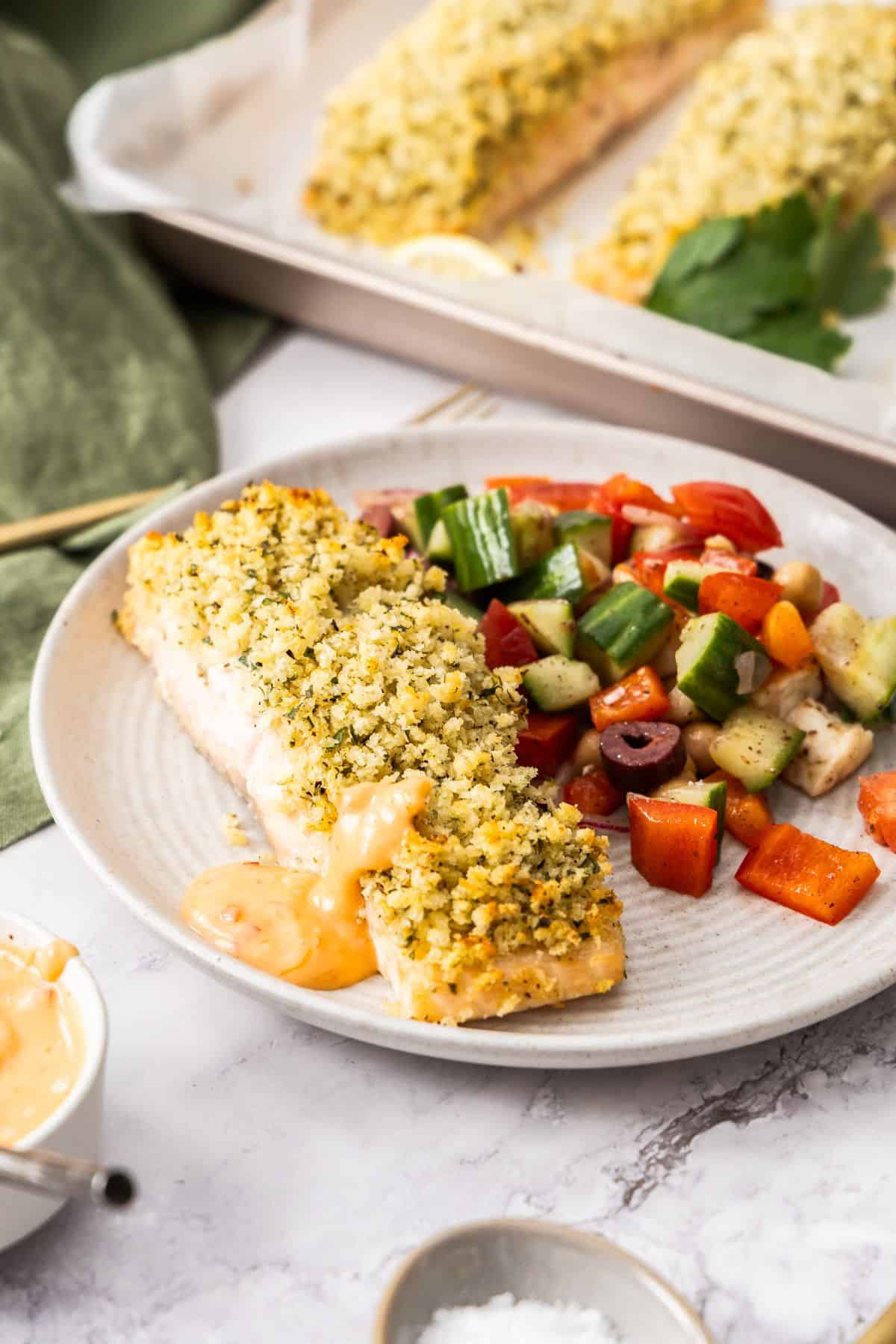 Round grey plate with a herb crusted salmon fillet, a drizzle of spicy mayo and a tomato salad on the side.