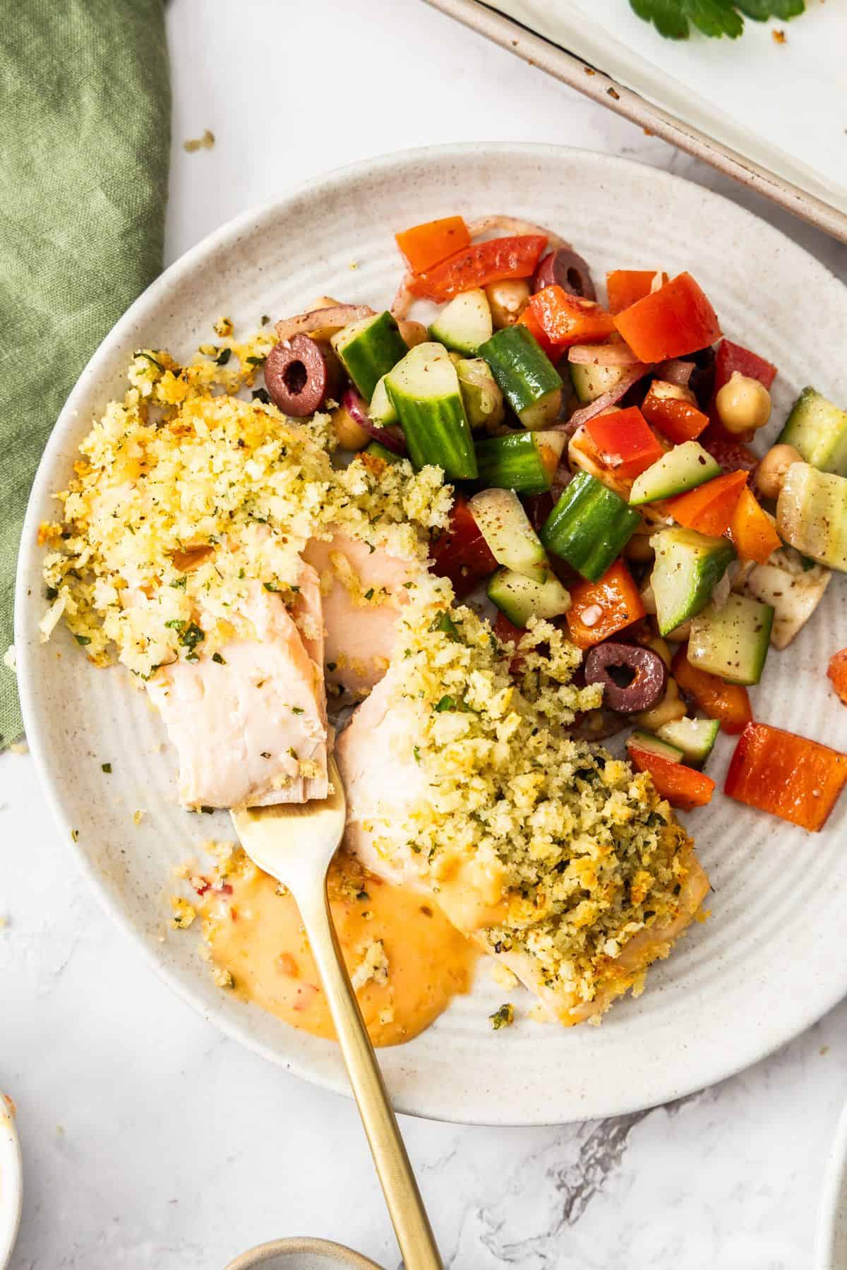 Fork cutting in to a piece of cooked salmon, sitting on a round grey plate, with a tomato salad and drizzle of spicy mayo.