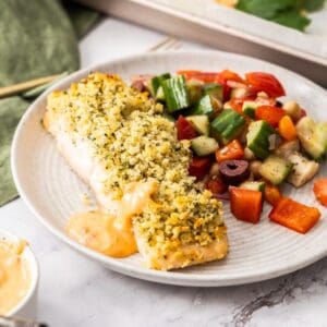 Round grey plate with a herb crusted salmon fillet, a drizzle of spicy mayo and a tomato salad on the side.