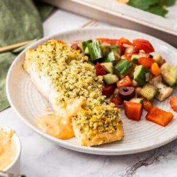 Round grey plate with a herb crusted salmon fillet, a drizzle of spicy mayo and a tomato salad on the side.