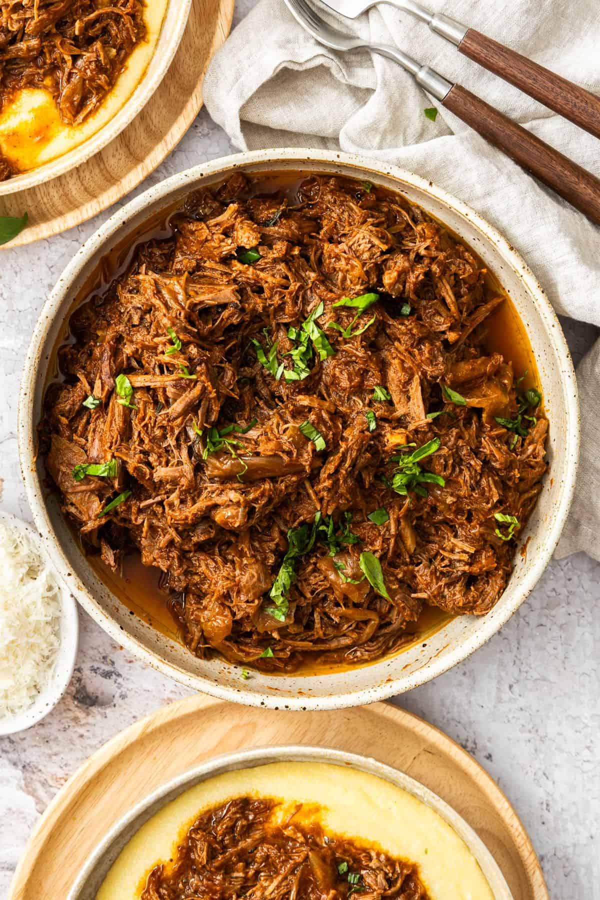 Round dish of Mediterranean Pulled Beef garnished with fresh basil, sitting on a white cloth.