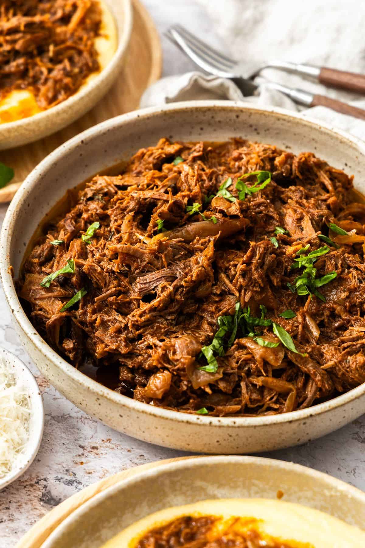 Round dish of Mediterranean Pulled Beef garnished with fresh basil, sitting on a white cloth.