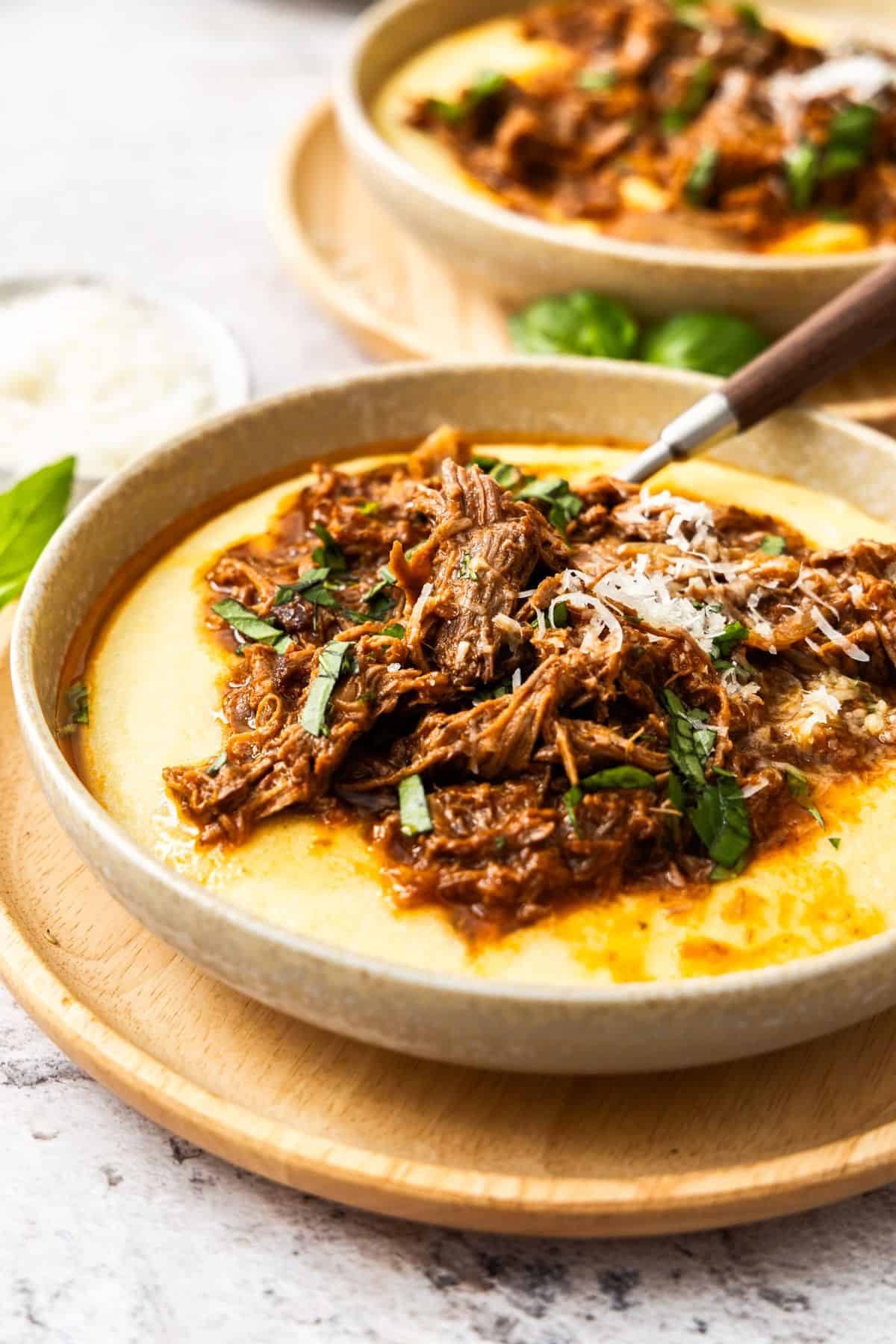 Round dish of Mediterranean Pulled Beef, on a serve of creamy polenta, on a round wooden mat, with a spoon in bowl.
