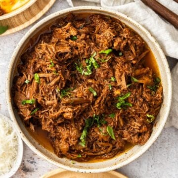 Round dish of Mediterranean Pulled Beef garnished with fresh basil, sitting on a white cloth.