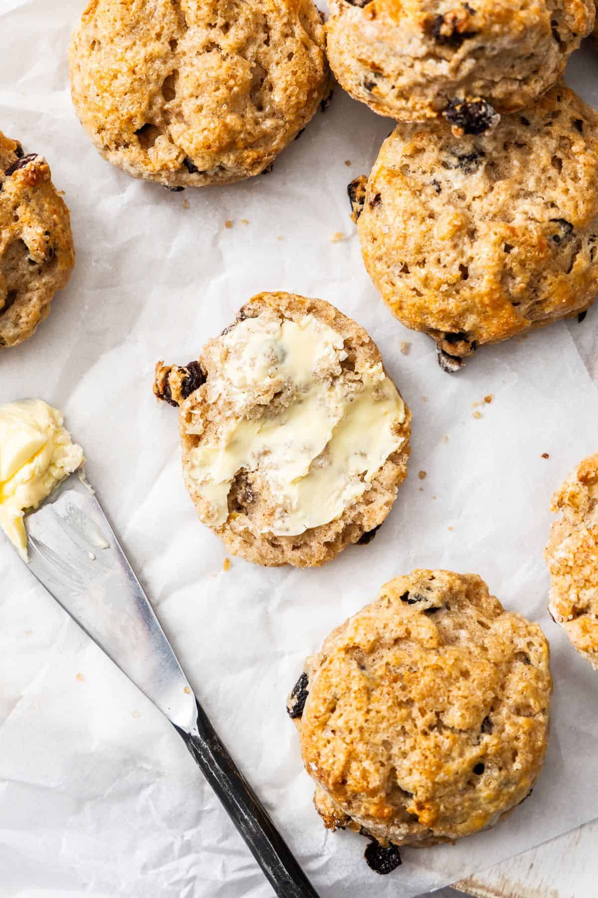 Freshly baked Sultana Scones stacked on some baking paper, with one cut in half spread with butter.
