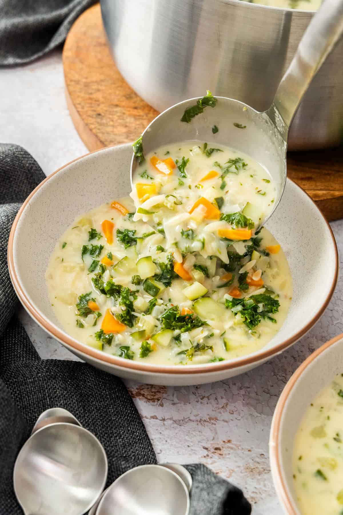 A soup ladle serving some Creamy Orzo Vegetable Soup into a round grey bowl.
