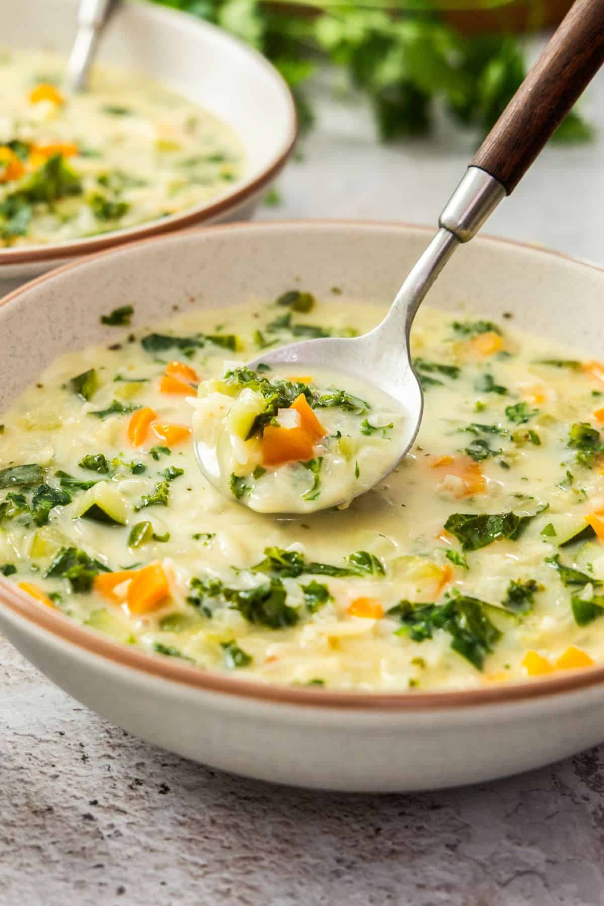 Round grey bowl with a serving of Creamy Orzo Vegetable Soup, with a spoon lifting up some soup.