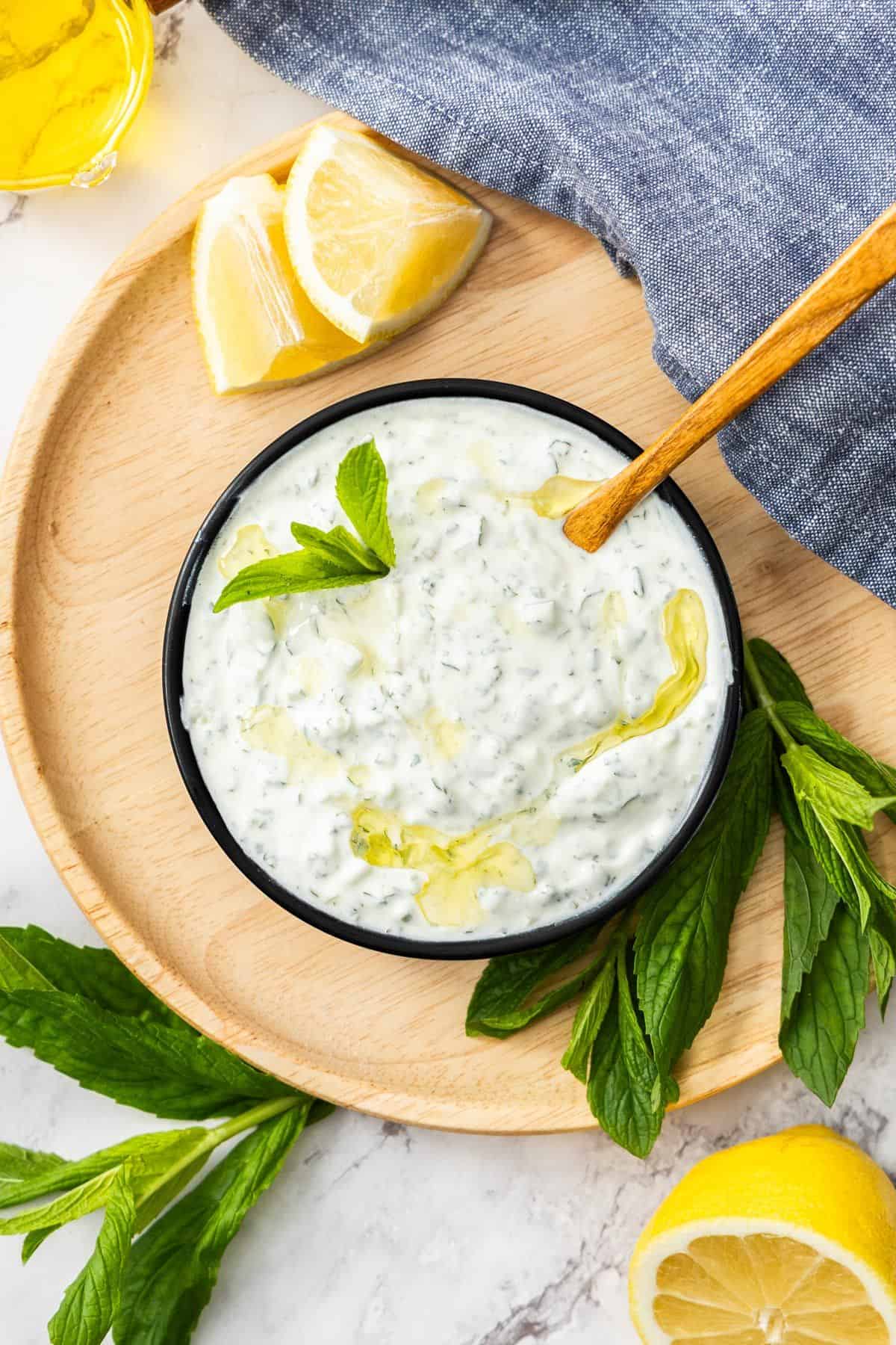 Small black dish sitting on wooden board of Cucumber and Yoghurt Dip, with a spoon resting in bowl.