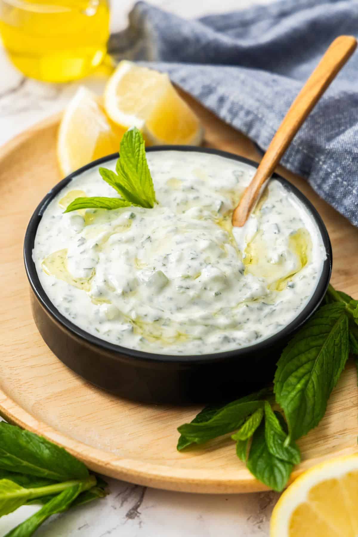 Small black dish sitting on wooden board of Cucumber and Yoghurt Dip, with a spoon resting in bowl.