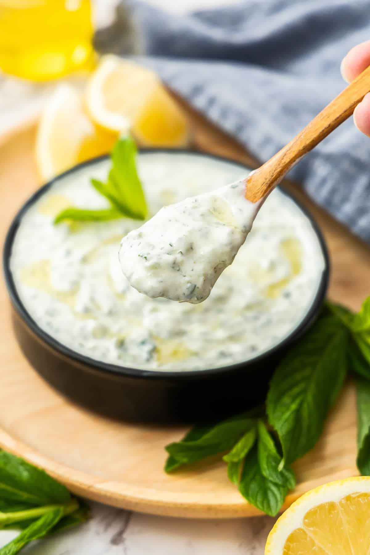 Small black dish sitting on wooden board of Cucumber and Yoghurt Dip, with a spoon lifting up some dip.